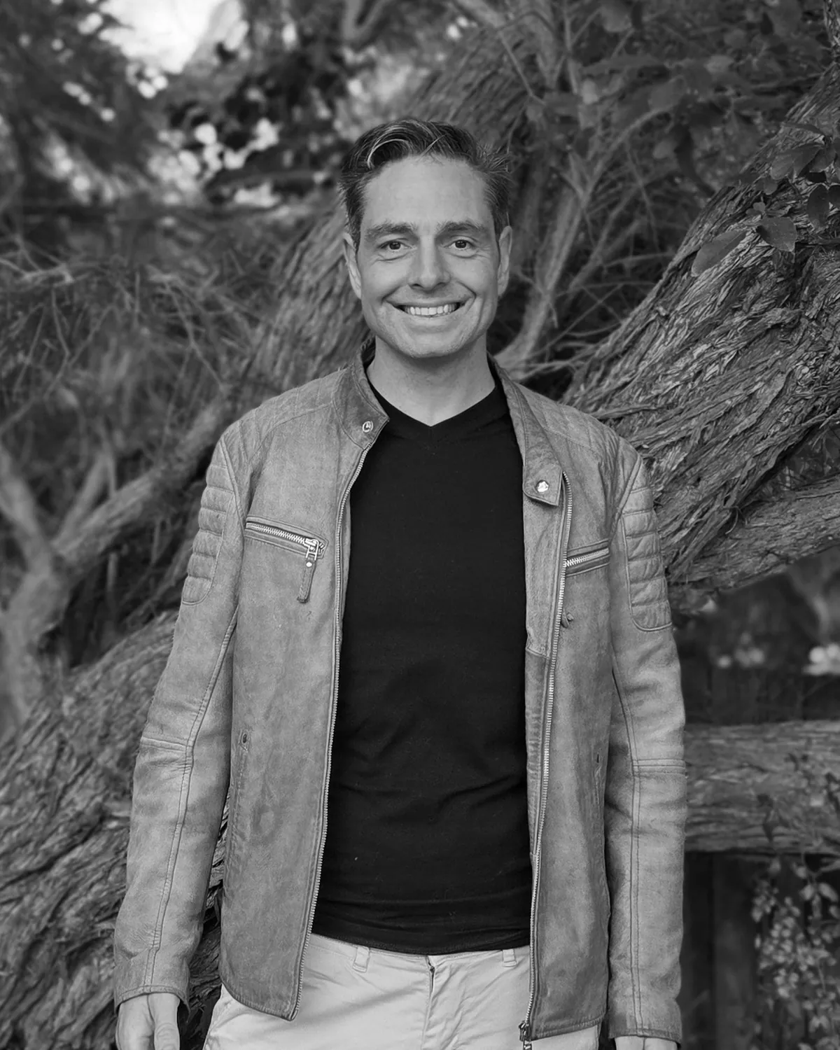 David Joshua smiling at the camera, in a leather jacket over a dark T-shirt, in front of a gnarled tree